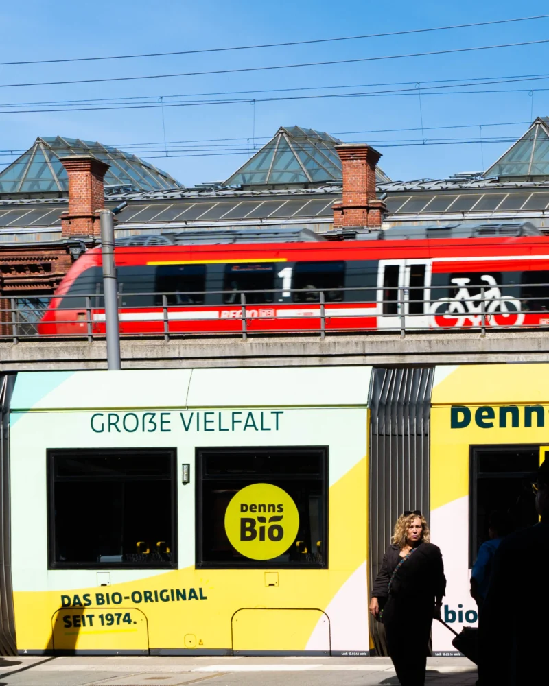 Alice · 03/05/2026 · 2165 x 2706 Cette photographie montre un tram jaune avec des inscriptions en allemand, notamment "Große Vielfalt" et "Bio-Original", promouvant des produits bio. Le sujet principal est ce tram, partiellement caché par un piéton.La composition met en valeur le tram et son message écologique. Le cadrage est serré sur le tram et inclut une partie du bâtiment et du ciel. La lumière est naturelle, avec un éclairage doux et des ombres nettes. Les couleurs dominantes sont le jaune vif du tram, contrastant avec le rouge du toit et le bleu du ciel.L’ambiance est urbaine et moderne, avec une touche de promotion environnementale.