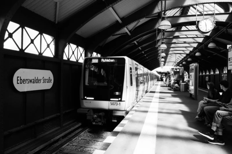 Alice · 03/05/2026 · 7008 x 4672 Cette photographie en noir et blanc montre un train de métro à l'arrêt à la station "Eberwacher Straße". Le sujet principal est le train moderne, bien éclairé, avec des passagers à l'intérieur et sur le quai.La composition met en valeur la symétrie des rails et des quais, avec une perspective légèrement en contre-plongée. Le cadrage est serré, capturant l’essentiel de la scène.La lumière est naturelle et filtrée, créant des contrastes doux. Les couleurs dominantes sont les tons de gris, renforçant l’ambiance urbaine et industrielle. Le style est documentaire, évoquant une atmosphère calme et quotidienne.