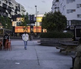 Alice · 07/05/2026 · 5726 x 3817 Cette photographie montre un homme marchant seul au centre d'une place urbaine. La composition met en valeur une perspective large avec des bâtiments modernes en arrière-plan et des éléments architecturaux géométriques. Le cadrage est légèrement bas, créant une impression de profondeur.La lumière est douce et tamisée, typique d’un crépuscule ou d’un temps nuageux. Les couleurs dominantes sont des tons neutres (gris, beige) avec des touches de vert et de bleu. Le style est réaliste et documentaire, évoquant une ambiance calme et légèrement mélancolique.