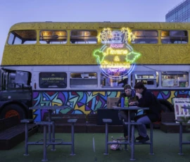 Alice · 07/05/2026 · 5992 x 3995 Cette photographie met en scène un bus à impériale décoré de motifs lumineux et colorés, principalement jaunes et verts. Le sujet principal est un personnage assis à une table devant le bus, éclairé par des néons.La composition joue sur un cadrage serré, mettant en avant les détails lumineux du bus et l’ambiance festive. Les couleurs dominantes sont vives et contrastées, avec des touches de bleu et de noir pour les contours.Le style évoque une ambiance urbaine et artistique, presque onirique, renforcée par les jeux de lumière et les motifs graphiques. L’ambiance est festive et créative, typique d’un événement nocturne ou d’une installation artistique.