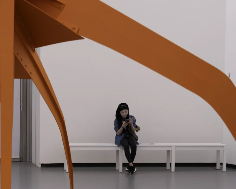 Alice · 23/05/2026 · 3556 x 2845 Cette photographie met en scène une femme assise sous une structure architecturale moderne en forme d’arche orange. Le sujet principal est cette femme, absorbée par son téléphone, créant un contraste entre son isolement et l’espace ouvert.La composition joue sur les lignes dynamiques de l’arche, encadrant la personne. Le cadrage est serré, centré sur elle et l’élément architectural. La lumière est douce et naturelle, éclairant uniformément la scène. Les couleurs dominantes sont le blanc des murs et l’orange vif de la structure, apportant une ambiance minimaliste et épurée. Le style est contemporain, évoquant une atmosphère calme et introspective.