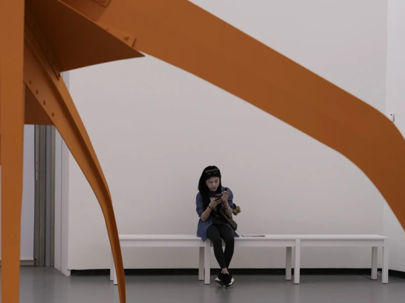 Cette photographie met en scène une femme assise sous une structure architecturale moderne en forme d’arche orange. Le sujet principal est cette femme, absorbée par son téléphone, créant un contraste entre son isolement et l’espace ouvert.La composition joue sur les lignes dynamiques de l’arche, encadrant la personne. Le cadrage est serré, centré sur elle et l’élément architectural. La lumière est douce et naturelle, éclairant uniformément la scène. Les couleurs dominantes sont le blanc des murs et l’orange vif de la structure, apportant une ambiance minimaliste et épurée. Le style est contemporain, évoquant une atmosphère calme et introspective.