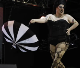 Alice · 24/05/2026 · 2017 x 2521 Cette photographie montre une personne en costume de scène excentrique, probablement un artiste de cabaret ou de burlesque. Le sujet principal est vêtu d’un costume moulant noir et or, avec une perruque extravagante et des accessoires colorés.La composition met en valeur un jeu de lumière dynamique, avec un éclairage théâtral qui accentue les contours. Le cadrage serré isole le sujet, créant une ambiance intense et dramatique. Les couleurs dominantes sont le noir, l’or et des touches de blanc et rose. Le style évoque le spectacle et l’exubérance, renforcé par l’usage d’un éventail stylisé. L’ambiance est à la fois sensuelle et provocante.