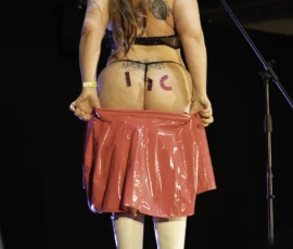 Alice · 24/05/2026 · 1678 x 2517 Cette photographie montre une personne sur scène, vue de dos, portant une tenue excentrique : un haut transparent révélant un faux ventre gonflé avec des lettres "LUC", une jupe rouge et des bottes blanches. Le cadrage est serré, centré sur elle, avec un fond flou de structure métallique.La lumière est vive et directionnelle, mettant en valeur les détails de la tenue. Les couleurs dominantes sont le rouge de la jupe, le blanc des bottes et le transparent du haut. Le style est provocateur et théâtral, créant une ambiance décalée et spectaculaire.