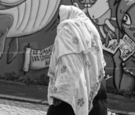 Pierre · 14/05/2026 · 5472 x 3080 Cette photographie en noir et blanc met en scène une personne vêtue d'un long manteau clair, marchant devant un mur tagué. Le sujet principal est flou, suggérant un mouvement.La composition est simple, avec un cadrage serré sur la silhouette et le graffiti coloré en arrière-plan. La lumière semble naturelle, douce, sans contraste marqué.Les couleurs dominantes, bien que réduites au noir et blanc, évoquent des tons clairs et sombres contrastés. Le style est réaliste et documentaire, capturant une scène urbaine. L'ambiance est à la fois mystérieuse et dynamique, reflétant une atmosphère de rue animée.