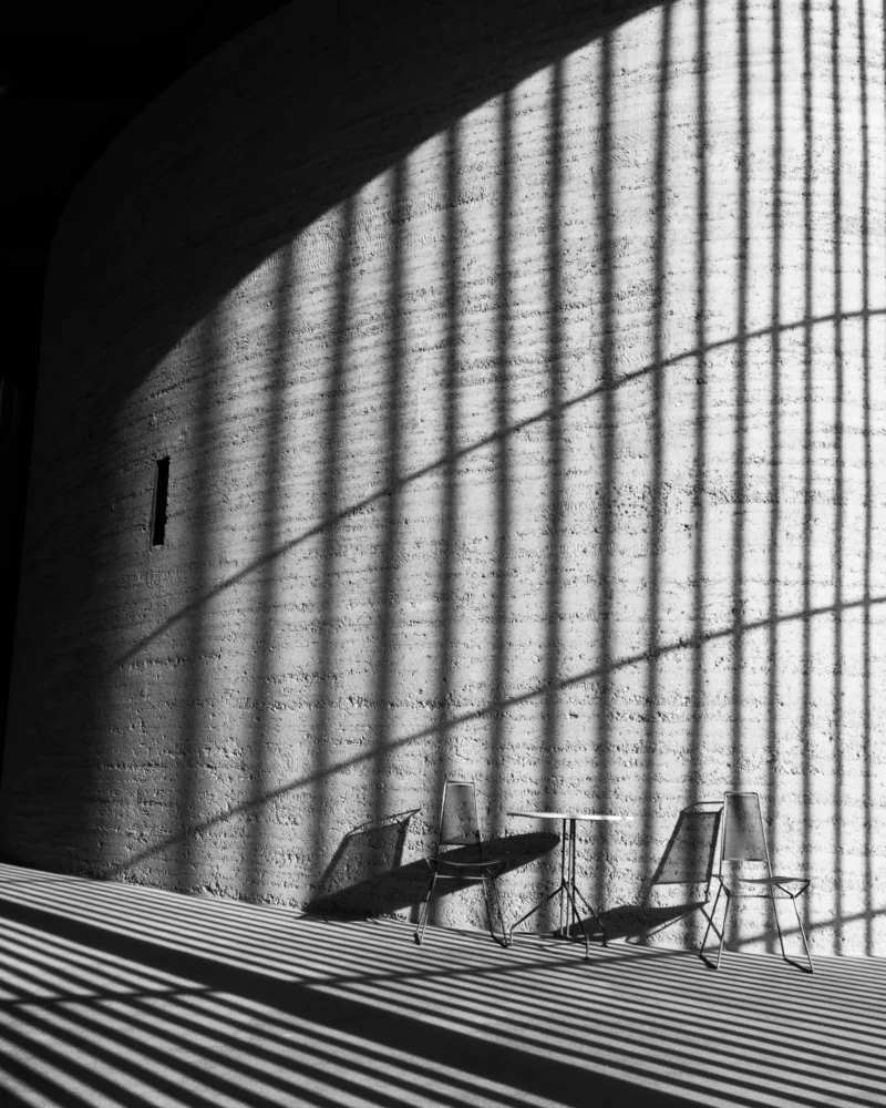 Alice · 30/04/2026 · 4553 x 5691 Cette photographie en noir et blanc met en scène deux chaises vides placées sur une surface en bois striée, devant une façade architecturale moderne aux lignes horizontales marquées. Le sujet principal est l’espace vide entre les chaises et le bâtiment, soulignant une impression de solitude et de minimalisme.Le cadrage serré et la composition géométrique créent une ambiance contemplative. La lumière naturelle, filtrée et contrastée, accentue les ombres et les textures. Les tons dominants, du blanc au noir en passant par les gris, renforcent le style épuré et intemporel, évoquant une atmosphère à la fois moderne et mélancolique.
