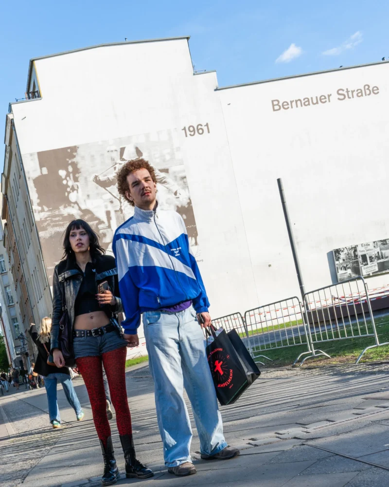 Alice · 30/04/2026 · 2753 x 3441 Cette photographie montre deux jeunes gens posant devant un bâtiment marqué "Bernauer Straße 1961". Le sujet principal est le couple, avec une composition centrée sur eux.Le cadrage est serré sur les personnages, avec un arrière-plan architectural partiellement visible. La lumière est naturelle et éclatante, suggérant un jour ensoleillé. Les couleurs dominantes sont les tons froids (bleu du manteau, gris du bâtiment) contrastant avec les teintes chaudes (peau, rouge des vêtements de la femme).Le style est documentaire et spontané, créant une ambiance moderne et urbaine.