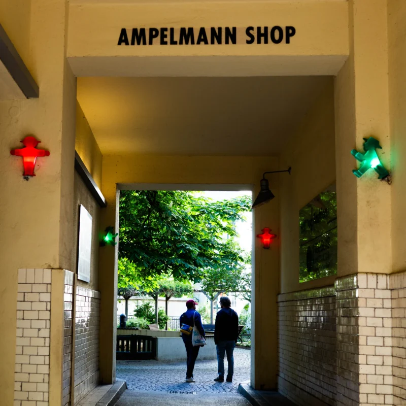 Alice · 01/05/2026 · 3009 x 3009 Cette photographie montre l'entrée de l’**Ampelmann Shop**, avec deux personnes marchant sous une arche. Le **sujet principal** est l’entrée du magasin, encadrée par des **lumières rouges et vertes** en forme de croix.La **composition** est symétrique, avec un cadrage serré sur l’arche. La **lumière** est naturelle et tamisée, renforcée par les éclairages artificiels. Les **couleurs dominantes** sont les tons beiges, verts et les éclats rouges/verts des lumières.Le style est **urbain et minimaliste**, évoquant une ambiance **moderne et légèrement industrielle**, avec une touche de nostalgie.
