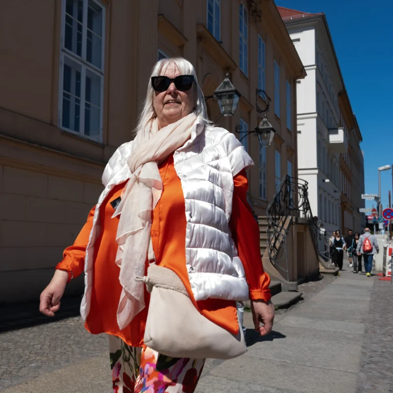 Alice · 01/05/2026 · 4672 x 4672 Cette photographie met en scène une personne âgée comme sujet principal, vêtue d’un style excentrique et coloré. Elle porte un manteau blanc et orange, des lunettes noires et des gants, avec une écharpe et des accessoires voyants.La composition est dynamique, avec un cadrage serré sur le sujet en mouvement. L’arrière-plan montre une rue urbaine avec des bâtiments classiques et quelques passants.La lumière est naturelle et vive, suggérant un jour ensoleillé. Les couleurs dominantes sont les tons chauds (orange, blanc) et quelques touches pastel. L’ambiance est à la fois décalée et joyeuse, mêlant originalité et quotidien.