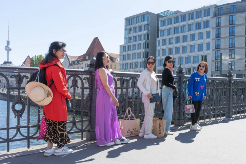Alice · 01/05/2026 · 5362 x 3575 Cette photographie montre un groupe de cinq personnes posant sur un pont en ville. Le sujet principal est leur diversité vestimentaire : robes colorées (rouge, violet) et tenues modernes (jeans, baskets).La composition est symétrique, avec les personnes alignées sur une balustrade métallique. Le cadrage inclut un arrière-plan urbain avec des bâtiments et un ciel dégagé. La lumière est naturelle et vive, suggérant un jour ensoleillé. Les couleurs dominantes sont vives et contrastées : rouge, violet, bleu, blanc et gris.Le style est documentaire et spontané, créant une ambiance joyeuse et décontractée.