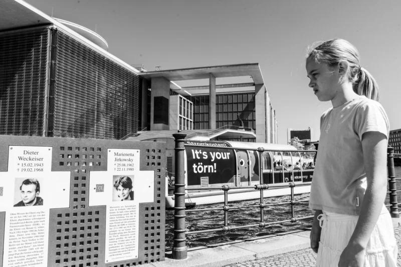 Alice · 01/05/2026 · 7008 x 4672 Cette photographie en noir et blanc montre une jeune fille comme sujet principal, observant des panneaux d'information devant une structure métallique.La composition met en avant les affiches avec des portraits et des textes, dont une question "It's your turn?" La lumière est naturelle et semble provenir d'un angle élevé, créant des contrastes nets. Le cadrage est serré sur la jeune fille et les panneaux, avec un arrière-plan flou.L'ambiance est sobre et réfléchie, renforcée par le monochrome, évoquant une visite éducative ou commémorative.