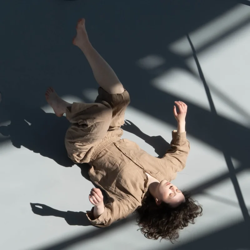Alice · 01/05/2026 · 1520 x 1520 Cette photographie capture une personne en mouvement, effectuant une roulade ou un mouvement acrobatique sur un sol clair. Le sujet principal est flou, suggérant dynamisme.La composition est épurée, centrée sur la silhouette. Le cadrage serré isole le sujet, mettant en valeur ses membres en extension. La lumière naturelle, probablement solaire, crée des ombres nettes et contrastées, accentuant les contours.Les couleurs dominantes sont les tons neutres (beige, gris) avec des contrastes forts entre lumière et ombre. Le style est documentaire et spontané, évoquant une ambiance à la fois ludique et artistique, pleine d'énergie et de liberté.