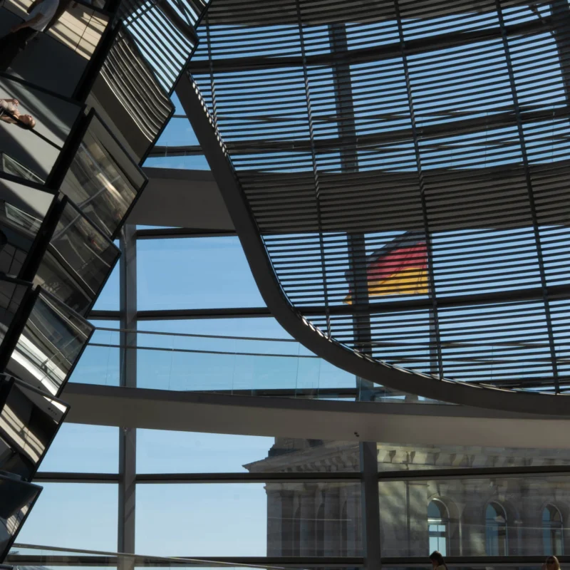 Alice · 01/05/2026 · 2293 x 2293 Cette photographie met en scène le **dôme en verre du Reichstag** à Berlin. Le **sujet principal** est le drapeau allemand, visible à travers la structure transparente. La **composition** joue sur les lignes géométriques des poutres métalliques et la transparence du verre, créant un effet de profondeur.Le **cadrage** est serré, centré sur le drapeau et les éléments architecturaux. La **lumière** naturelle, vive et contrastée, accentue les reflets et les ombres. Les **couleurs dominantes** sont le bleu du ciel, le gris métallique et les teintes rouges, noires et jaunes du drapeau.Le style est **moderne et épuré**, évoquant une ambiance de **transparence politique et d’ouverture**.
