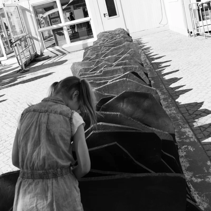 Alice · 01/05/2026 · 4672 x 4672 Cette photographie en noir et blanc montre une personne, vue de dos, penchée sur une structure architecturale géométrique et allongée, évoquant une sculpture ou un élément décoratif. Le sujet principal est cette interaction entre l’humain et l’objet.La composition joue sur les lignes droites et les angles aigus de la structure, créant un contraste avec la posture courbée de la personne. Le cadrage est serré, centré sur cette scène. La lumière semble naturelle, avec des ombres marquées, renforçant le côté dramatique. L’absence de couleurs accentue les textures et les contrastes lumineux, donnant une ambiance à la fois contemplative et mystérieuse. Le style rappelle le photojournalisme ou une approche documentaire.