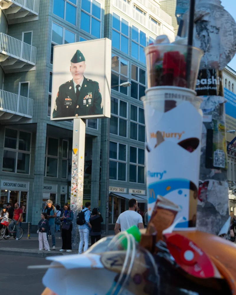 Alice · 01/05/2026 · 2249 x 2811 Cette photographie montre une manifestation ou un hommage. Le sujet principal est une bannière avec le portrait d’un homme en uniforme militaire, entourée de bougies et d’objets divers (vêtements, jouets, fleurs).La composition est serrée, avec un cadrage en plan rapproché sur les objets devant. La lumière est naturelle, probablement en journée, avec des ombres marquées. Les couleurs dominantes sont sobres : gris des bâtiments, noir des bougies, et touches de rouge et blanc sur les objets.L’ambiance est solennelle et émouvante, évoquant un hommage ou une commémoration. Le style est documentaire et spontané.