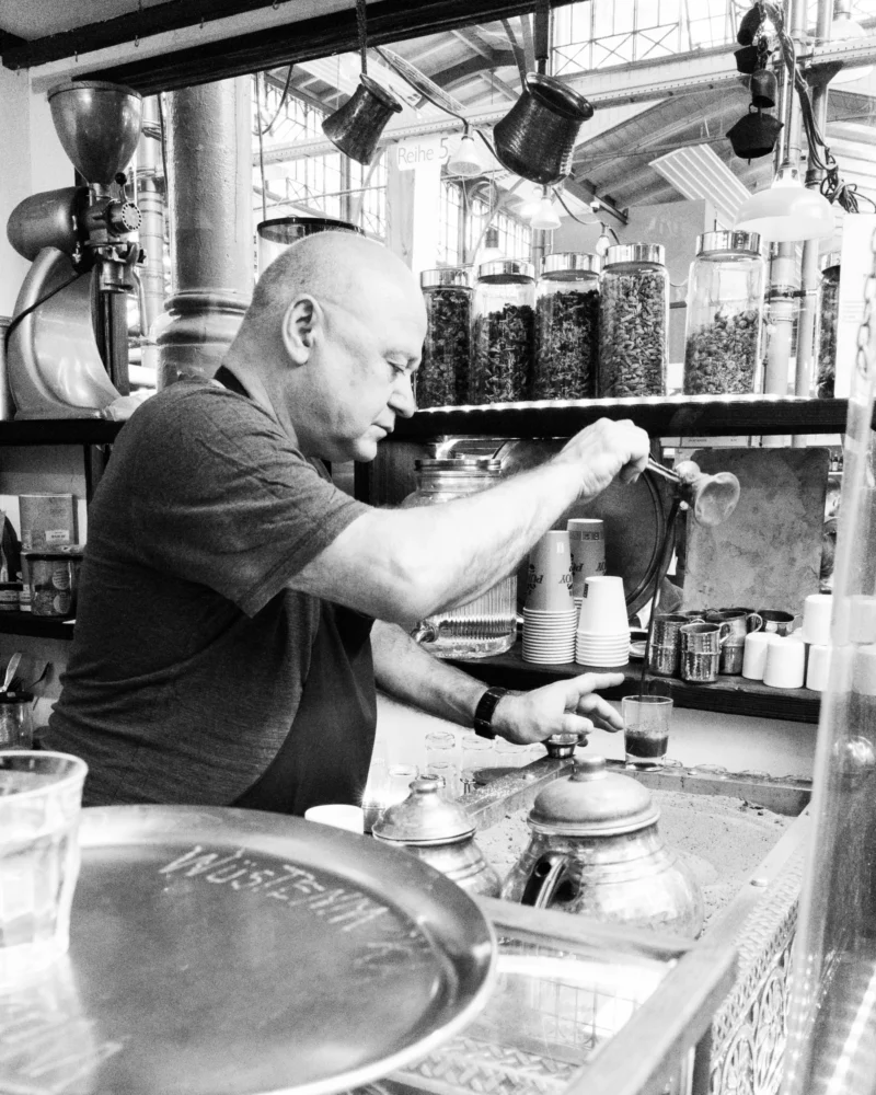 Alice · 02/05/2026 · 4410 x 5512 Cette photographie en noir et blanc montre un homme comme sujet principal, probablement un barista, préparant des boissons derrière un comptoir.La composition met en valeur son activité, avec des éléments comme des machines à café et des accessoires en arrière-plan. Le cadrage est serré, centré sur ses gestes précis.La lumière est naturelle et douce, éclairant bien le sujet sans créer d'ombres marquées. L'absence de couleurs renforce l'atmosphère intemporelle et artisanale.Le style est documentaire, authentique, évoquant une ambiance chaleureuse et professionnelle dans un cadre typique de café.