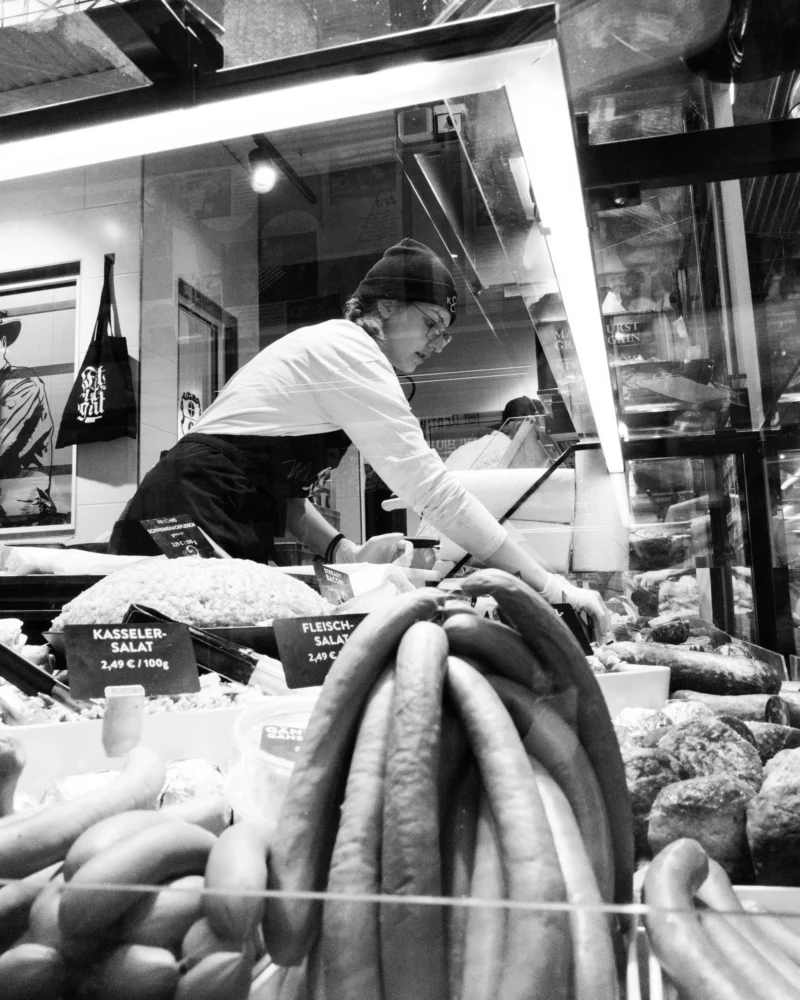 Alice · 02/05/2026 · 4672 x 5840 Cette photographie en noir et blanc montre une personne, probablement un boucher, travaillant derrière un comptoir de viande. Le sujet principal est centré sur cette personne en action, entourée de viandes exposées.La composition est dynamique, avec un cadrage serré sur le comptoir et le sujet. La lumière est naturelle et met en valeur les détails des produits et des outils. L’ambiance est artisanale et authentique, évoquant un marché ou une boucherie traditionnelle. Le style est documentaire, captant un moment réaliste et quotidien. Les tons clairs et ombres renforcent l’aspect rétro et brut de la scène.