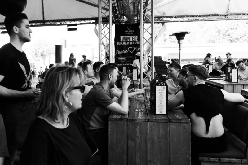 Alice · 02/05/2026 · 5085 x 3390 Cette photographie en noir et blanc capture une scène de terrasse animée. Le sujet principal est un groupe de personnes assises à des tables, semblant discuter ou attendre. La composition montre une profondeur avec des plans superposés, des gens en arrière-plan légèrement flous.Le cadrage est serré sur les visages et les épaules des individus au premier plan. La lumière est douce, avec des contrastes modérés, créant une ambiance chaleureuse et détendue. Les couleurs, bien que monochromes, suggèrent une ambiance nocturne ou crépusculaire.Le style est documentaire et spontané, évoquant une ambiance conviviale et décontractée.