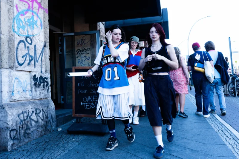 Alice · 02/05/2026 · 5299 x 3533 Cette photographie montre deux personnes en costume déguisées, probablement pour une parade ou un événement festif. Le sujet principal est l’homme en tenue de footballeur vintage (numéro 01) et la femme en robe blanche.La composition met en avant leur interaction devant une porte ornée de graffitis. Le cadrage est serré sur eux, avec d’autres participants flous en arrière-plan. La lumière est naturelle, légèrement tamisée, créant des ombres douces. Les couleurs dominantes sont les tons pastel (blanc, bleu) et les contrastes urbains (noir des graffitis, gris de la façade).L’ambiance est festive et décontractée, dans un style streetwear et artistique.