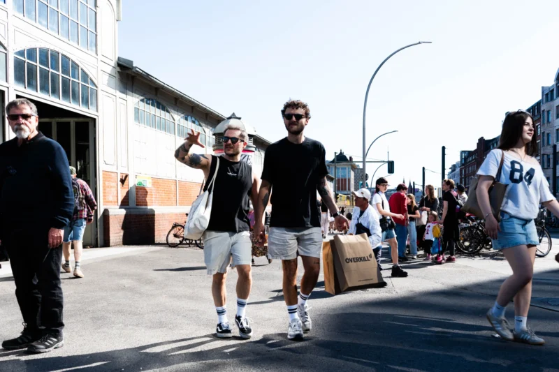 Alice · 02/05/2026 · 5611 x 3741 Cette photographie capture une scène urbaine animée. Le sujet principal semble être un groupe de jeunes hommes en mouvement, dont l’un porte un chapeau et un short, tenant un objet en forme de raquette ou de planche.La composition est dynamique, avec un cadrage légèrement bas, mettant en valeur les personnages en premier plan. L’ambiance est décontractée et festive, avec une foule en arrière-plan.La lumière est naturelle et abondante, suggérant un jour ensoleillé. Les couleurs dominantes sont claires et vives : tons chauds (beige, blanc) et contrastes avec des vêtements colorés. Le style est documentaire et spontané, reflétant une ambiance de vie quotidienne en extérieur.
