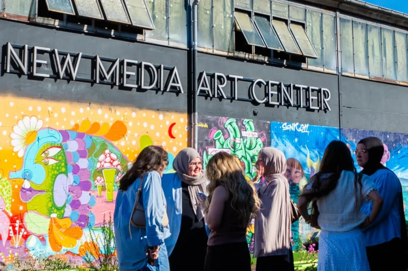 Alice · 02/05/2026 · 2107 x 1405 Cette photographie montre un groupe de personnes observant une fresque murale colorée devant le **New Media Art Center**. Le sujet principal est l’interaction sociale autour de l’art.La composition est dynamique, avec un cadrage serré sur les visiteurs et le mur. La lumière est naturelle et douce, mettant en valeur les couleurs vives de la fresque (bleus, roses, jaunes). L’ambiance est conviviale et artistique, reflétant un lieu culturel et créatif. Le style est réaliste et spontané, captant un moment authentique.
