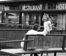 Dominique · 23/05/2026 · 1500 x 1000 Cette photographie en noir et blanc montre deux personnes assises sur un banc devant un établissement affichant "Bar", "Restaurant" et "Hôtel". Le sujet principal est la femme en robe légère, souriante, tenant la main de l’homme.La composition est symétrique, avec un cadrage serré sur les personnages. La lumière est douce et naturelle, créant des contrastes modérés. L’ambiance est chaleureuse et intimiste, évoquant une scène de détente ou de rencontre. Le style est documentaire, avec un rendu réaliste et une touche nostalgique grâce au noir et blanc.