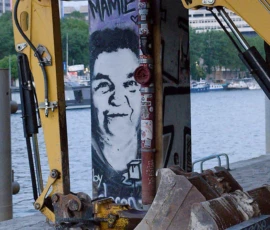 Dominique · 23/05/2026 · 667 x 1000 Cette photographie met en scène un **excavateur Yanmar** devant un **muret tagué**. Le sujet principal est le **visage souriant stylisé** peint en noir et rouge sur le mur.La **composition** joue sur le contraste entre la machine industrielle et l’art urbain. Le **cadrage** est serré, centré sur le graffiti et partiellement sur l’engin. La **lumière** semble naturelle, avec des ombres marquées.Les **couleurs dominantes** sont le jaune vif de l’excavateur, le noir et rouge du graffiti, et les tons gris du mur et de l’eau en arrière-plan. Le style est **urbain et contrasté**, créant une ambiance à la fois **industrielle et artistique**.