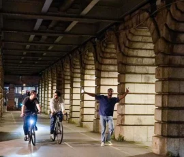 Dominique · 23/05/2026 · 667 x 1000 Cette photographie montre un tunnel urbain éclairé où des cyclistes et des piétons se déplacent. Le sujet principal est un groupe de personnes, dont un homme en bleu sur un vélo en premier plan.La composition est dynamique, avec une perspective en profondeur accentuée par les colonnes et les jeux d’ombres. Le cadrage est serré mais laisse voir l’espace autour.La lumière est artificielle, douce et légèrement bleutée, créant une ambiance nocturne ou crépusculaire. Les couleurs dominantes sont les tons gris et bleus, avec des éclats de vêtements colorés.Le style est réaliste et documentaire, évoquant une ambiance moderne et urbaine.