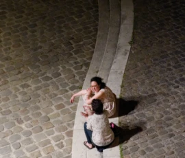 Dominique · 23/05/2026 · 15459 x 23184 Cette photographie montre deux enfants assis sur une bordure en pierre courbe, l’un tenant l’autre. Le sujet principal est leur interaction, évoquant une complicité ou un jeu.La composition est dynamique, avec une perspective plongeante accentuée par le cadrage serré. La lumière, tamisée et contrastée, crée des ombres marquées, soulignant les contours. Les couleurs dominantes sont les tons froids (gris des pavés, blanc de la bordure) et quelques touches chaudes (vêtements des enfants).Le style est documentaire et naturel, capturant un moment spontané. L’ambiance est à la fois intimiste et légèrement mystérieuse, renforcée par la nuit et les jeux d’ombres.