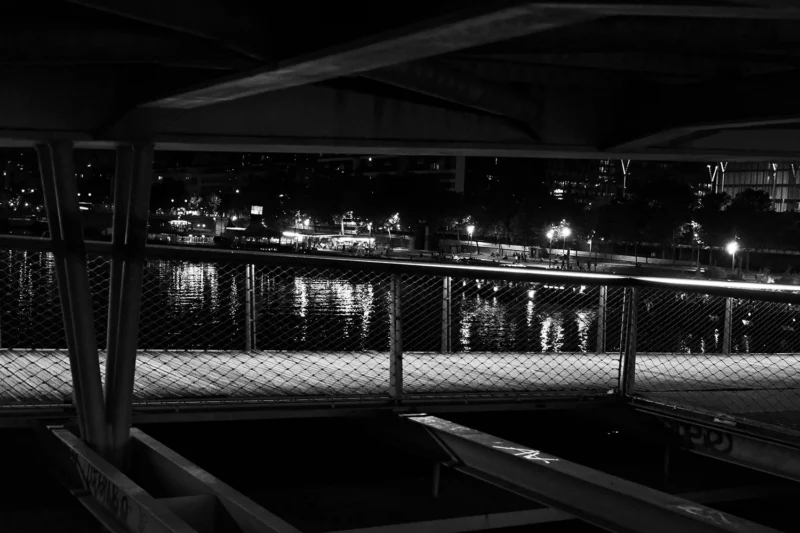 Dominique · 24/05/2026 · 1500 x 999 Cette photographie en noir et blanc capture une scène nocturne urbaine vue depuis une passerelle ou un pont. Le sujet principal est une ville illuminée reflétée dans l’eau calme d’un plan d’eau en contrebas.La composition repose sur des lignes horizontales et verticales, encadrées par des garde-corps métalliques. Le cadrage est serré, mettant en valeur les lumières urbaines et leur reflet. Les couleurs dominantes sont les nuances de gris, accentuées par les éclairages jaunes et blancs.Le style est épuré et contemplatif, évoquant une ambiance paisible et mélancolique, presque onirique.
