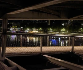 Dominique · 24/05/2026 · 1500 x 1000 Cette photographie capture une scène nocturne depuis un pont ou une passerelle. Le sujet principal est une vue urbaine sur une rivière ou un lac, avec des lumières distantes reflétées sur l’eau.La composition est équilibrée, avec une barrière métallique en premier plan encadrant la vue. Le cadrage est large, incluant des éléments architecturaux comme des ponts et des bâtiments éclairés. La lumière est tamisée, avec des teintes bleutées et des éclairages artificiels créant une ambiance nocturne mystérieuse. Les couleurs dominantes sont les noirs, gris et bleus, renforçant une atmosphère calme et contemplative. Le style est réaliste et photographique, évoquant une ambiance sereine et presque mélancolique.