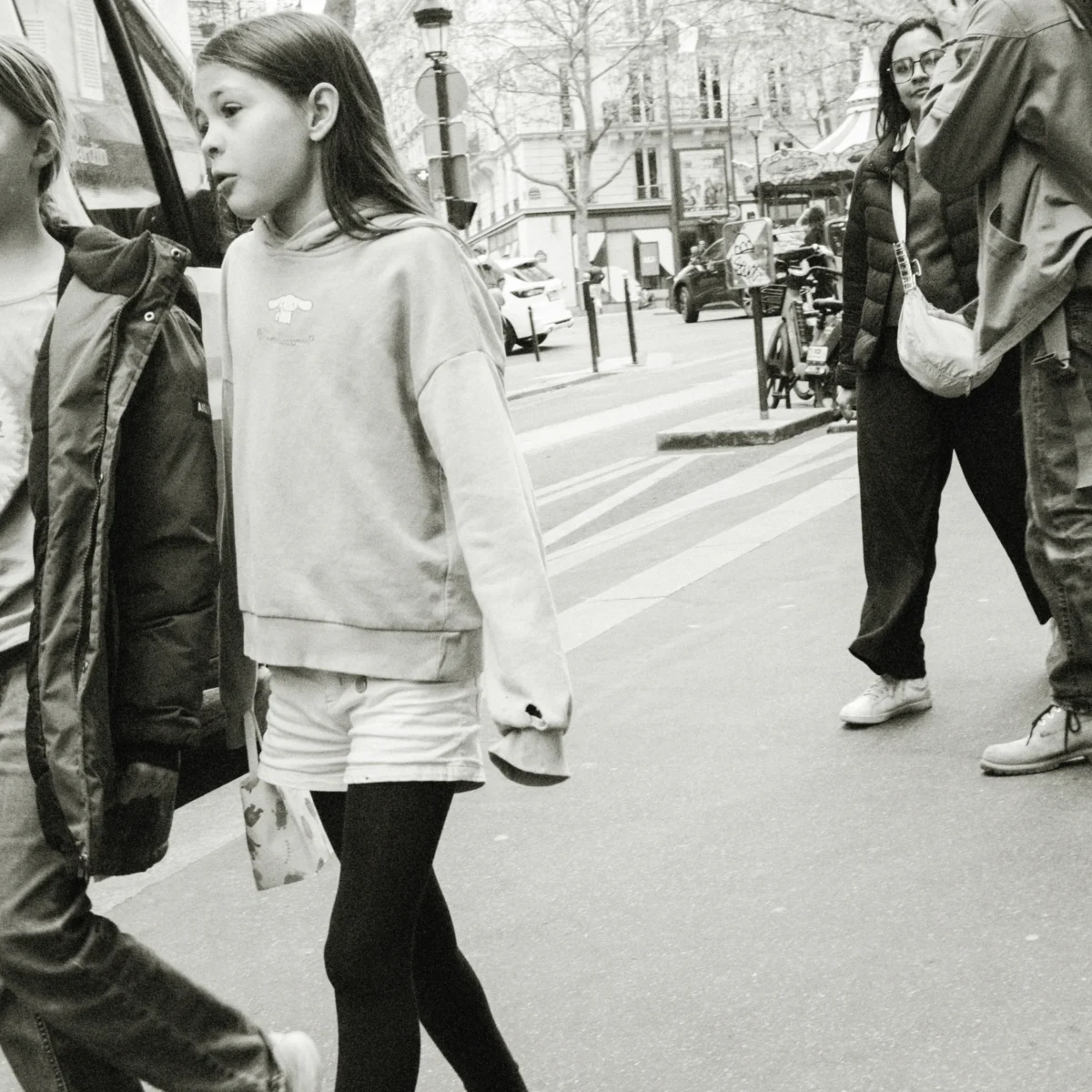 Nathalie F. · 11/04/2026 · 4160 x 4160 Cette photographie en noir et blanc met en scène une jeune fille comme sujet principal, marchant au centre de l’image. La composition montre une rue urbaine avec des passants en arrière-plan.Le cadrage est serré sur elle, capturant son mouvement et son expression. La lumière est naturelle, avec des contrastes marqués créant des ombres nettes. Les couleurs dominantes, bien que monochromes, varient des tons clairs aux noirs profonds.Le style est documentaire et spontané, évoquant une ambiance authentique et quotidienne, presque mélancolique. L’image capte une scène de vie urbaine, simple et intime.