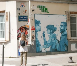 Nathalie F. · 09/05/2026 · 3678 x 3607 Cette photographie montre une femme en train de marcher devant un mur mural. Le **sujet principal** est le contraste entre elle et le **mural coloré** derrière, représentant des personnages stylisés en bleu.La **composition** est équilibrée, avec un **cadrage serré** sur la femme et le mur. La **lumière** naturelle éclaire la scène, créant des ombres douces. Les **couleurs dominantes** sont les tons froids du bleu du mural et les teintes neutres de la façade.Le **style** est urbain et artistique, mêlant street art et vie quotidienne. L’**ambiance** est dynamique et vivante, reflétant une atmosphère moderne et créative.