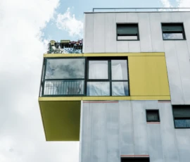 Nathalie F. · 09/05/2026 · 4160 x 4160 Cette photographie met en scène un bâtiment moderne avec une structure architecturale originale. Le sujet principal est une extension jaune en saillie, formant une sorte de balcon ou de volume suspendu.La composition joue sur les contrastes géométriques et les lignes verticales. Le cadrage est serré, centré sur cette partie saillante. La lumière naturelle, douce et légèrement diffuse, accentue les textures et les jeux d'ombres.Les couleurs dominantes sont le gris métallique du bâtiment et le jaune vif de l'extension, créant un style contemporain et audacieux. L'ambiance est à la fois minimaliste et futuriste, évoquant innovation et modernité.
