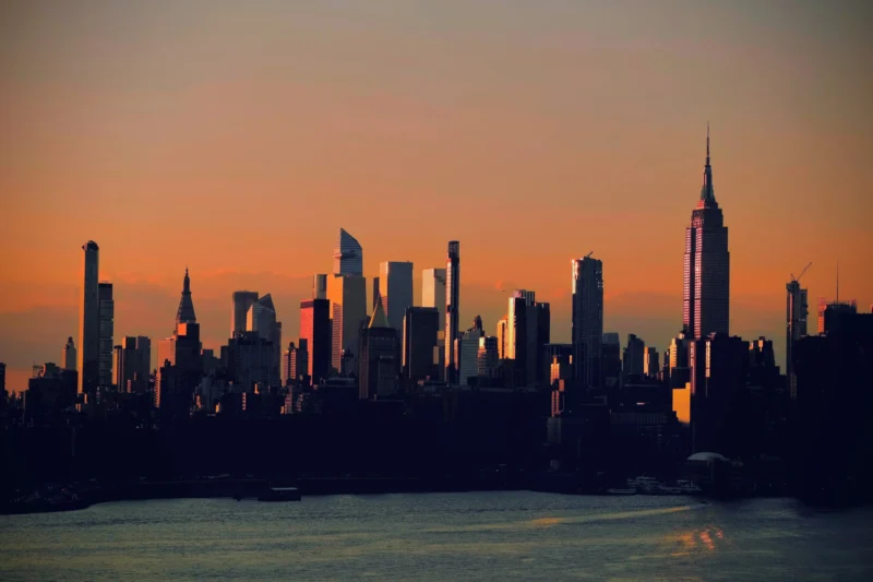Denis · 04/09/2025 · 4875 x 3250 Cette photographie capture un **skyline urbain au coucher de soleil**, probablement celui de New York. Le **sujet principal** est la silhouette des gratte-ciels, dont l’Empire State Building se détache. La **composition** est horizontale, avec une ligne d’horizon basse, mettant en valeur les bâtiments.Le **cadrage** est large, englobant une vue panoramique. La **lumière** douce et dorée crée un contraste entre les ombres des bâtiments et le ciel. Les **couleurs dominantes** sont des tons chauds (oranges, rouges) et des silhouettes sombres.Le **style** est réaliste et photographique, avec une **ambiance** à la fois sereine et majestueuse, évoquant la grandeur urbaine.