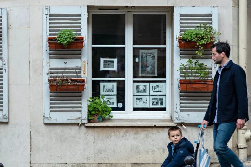 AnJo · 21/03/2026 · 1920 x 1280 Cette photographie met en scène deux hommes marchant côte à côte dans une rue urbaine. Le sujet principal est le jeu de contrastes entre eux et un bâtiment ancien aux fenêtres ornées de pots de fleurs.La composition est équilibrée, avec un cadrage serré sur les fenêtres et les plantes, créant une symétrie visuelle. La lumière naturelle éclaire la scène, douce et légèrement contrastée. Les couleurs dominantes sont les tons neutres (gris, beige) du bâtiment, adoucis par les touches de vert des plantes et les teintes bleues et grises des vêtements.Le style est réaliste et documentaire, capturant un instant de vie quotidienne. L’ambiance est calme et chaleureuse, mêlant modernité et charme vintage.