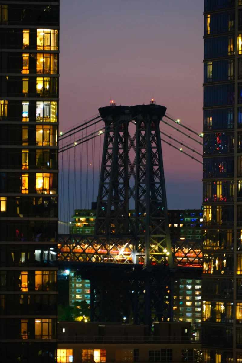 Denis · 04/09/2025 · 3265 x 4898 Cette photographie met en scène le **pont de Brooklyn** à New York, sujet principal. La composition est verticale, encadrée par des bâtiments modernes éclairés. Le cadrage isole le pont, avec une perspective basse, accentuant sa structure métallique.Les lumières, douces et artificielles, contrastent avec le ciel crépusculaire aux teintes **violet-bleutées**. Les couleurs dominantes sont les **tons froids** (bleu, violet) et les éclairages chauds (jaunes, blancs).Le style est **urbain et photographique**, avec une ambiance **sereine et légèrement mélancolique**, mêlant modernité et nostalgie.