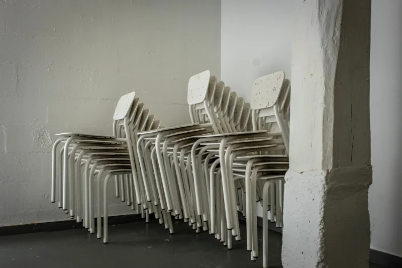 AnJo · 26/04/2026 · 1920 x 1280 Cette photographie montre une série de **chaises blanches en plastique** disposées en rangée contre un mur. Le **sujet principal** est ces chaises, alignées de manière uniforme.La **composition** est épurée, avec un **cadrage serré** mettant en valeur les chaises et le mur blanc. La **lumière** semble naturelle et douce, créant des ombres discrètes. Les **couleurs dominantes** sont le blanc des chaises et le gris clair du mur, renforçant une ambiance minimaliste.Le **style** est sobre et documentaire, évoquant une **ambiance froide et déserte**, presque industrielle ou scolaire.