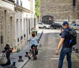 AnJo · 09/05/2026 · 1280 x 1920 Cette photographie capture une scène urbaine animée. Le sujet principal est un homme en jeans et veste bleue, debout avec un sac à dos, observant une femme sur un scooter électrique. Un photographe en arrière-plan immortalise la scène.La composition est dynamique, avec une perspective en contre-plongée le long d’une rue étroite bordée de bâtiments anciens. Le cadrage inclut des éléments architecturaux et des détails urbains.La lumière est naturelle et douce, typique d’une journée ensoleillée. Les couleurs dominantes sont les tons neutres des bâtiments, contrastés par les bleus et gris des vêtements. L’ambiance est à la fois quotidienne et artistique, mêlant mouvement et immobilité.