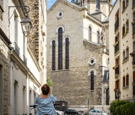 AnJo · 09/05/2026 · 1280 x 1920 Cette photographie met en scène un enfant assis sur un vélo dans une ruelle étroite. Le sujet principal est le bâtiment religieux imposant en arrière-plan, avec sa façade en pierre et ses grandes fenêtres.La composition joue sur la perspective avec un cadrage serré sur la ruelle menant vers l'édifice. La lumière est naturelle, douce et légèrement contrastée, créant des ombres marquées. Les couleurs dominantes sont les tons beiges et gris des bâtiments, adoucis par des touches de bleu dans le ciel.Le style est documentaire et pittoresque, évoquant une ambiance paisible et historique.
