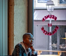 AnJo · 14/05/2026 · 1280 x 1920 Cette photographie met en scène un homme assis seul dans un café, absorbé par son téléphone. La composition joue sur la profondeur avec un cadrage serré sur le sujet, tout en laissant voir l'extérieur flou à travers la vitre.La lumière naturelle, douce et tamisée, crée une ambiance chaleureuse et intimiste. Les couleurs dominantes sont des tons neutres (beige, gris) rehaussés par des touches de rouge (enseigne "Restaurant") et de bois naturel.Le style est réaliste et documentaire, capturant un moment du quotidien. L’ambiance est calme et légèrement mélancolique, évoquant la solitude urbaine.