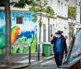 AnJo · 14/05/2026 · 1920 x 1280 Cette photographie met en scène une personne âgée, vêtue d’un manteau bleu, comme sujet principal, marchant devant un mur coloré. La composition associe un premier plan dynamique avec des éléments urbains (poubelles, trottoir) et un arrière-plan artistique, marqué par un mural représentant des oiseaux stylisés.Le cadrage est serré sur la personne, intégrant partiellement le mural. La lumière naturelle, douce et légèrement diffuse, éclaire la scène sans créer d’ombres marquées. Les couleurs dominantes sont vives (bleu, vert, jaune du mural) contrastant avec les tons neutres du bâtiment et du manteau.L’ambiance est à la fois urbaine et poétique, mêlant quotidien et art.