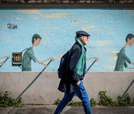 AnJo · 14/05/2026 · 1920 x 1280 Cette photographie montre un homme en mouvement, vêtu d’un manteau vert et d’un chapeau, marchant devant un mur mural. Le sujet principal est mis en valeur par un cadrage serré et une composition dynamique, avec des lignes directrices (le trottoir et les silhouettes murales).Les couleurs dominantes sont les bleus du mur, les verts du manteau et les gris de l’environnement urbain. La lumière semble naturelle, avec des contrastes modérés. Le style est réaliste et documentaire, créant une ambiance à la fois urbaine et artistique, mêlant vie quotidienne et art de rue.