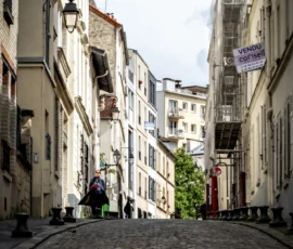 AnJo · 14/05/2026 · 1280 x 1920 Cette photographie capture une **ruelle pavée étroite** dans un quartier urbain européen. Le **sujet principal** est l'alignement des bâtiments anciens aux façades claires, encadrant la voie.La **composition** est symétrique, avec une perspective convergente vers l'arrière-plan. Le **cadrage** est serré, mettant en valeur les détails architecturaux. La **lumière** naturelle, douce et légèrement voilée, crée des ombres douces. Les **couleurs dominantes** sont les tons beiges, gris et verts, renforçant l'ambiance historique.Le **style** est réaliste et documentaire, évoquant une **ambiance paisible et nostalgique**, typique des vieilles villes.