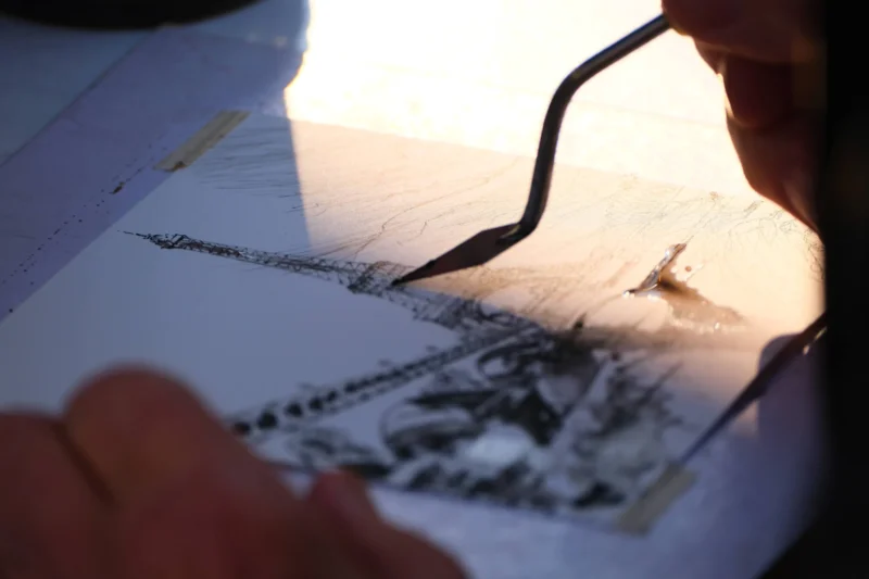 Denis · 21/03/2026 · 4898 x 3265 Cette photographie montre une personne traçant ou retouchant un dessin à l’aide d’un stylo-plume. Le sujet principal est le geste précis de la main tenant l’outil, sur un papier contenant une image en noir et blanc.La composition est serrée, centrée sur les détails du travail graphique. Le cadrage est rapproché, mettant en valeur les traits délicats. La lumière, douce et naturelle, éclaire le dessin depuis la gauche, créant des contrastes subtils. Les couleurs dominantes sont les tons gris et blancs, renforçant l’aspect artistique et minutieux.L’ambiance est studieuse et intime, évoquant concentration et patience.