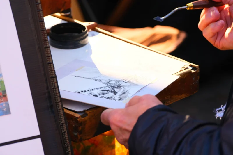 Denis · 21/03/2026 · 4898 x 3265 Cette photographie montre une personne en train de dessiner ou d'encrer une planche. Le sujet principal est les mains tenant un pinceau et un trait en cours d’exécution sur une feuille posée sur une planche à dessin.La composition est serrée, centrée sur le geste artistique. Le cadrage est rapproché, mettant en valeur les détails du travail. La lumière est naturelle et douce, éclairant le dessin et créant des contrastes. Les couleurs dominantes sont neutres, avec des tons gris et noirs, typiques d’un dessin à l’encre.L’ambiance est studieuse et artistique, évoquant la concentration et la précision.