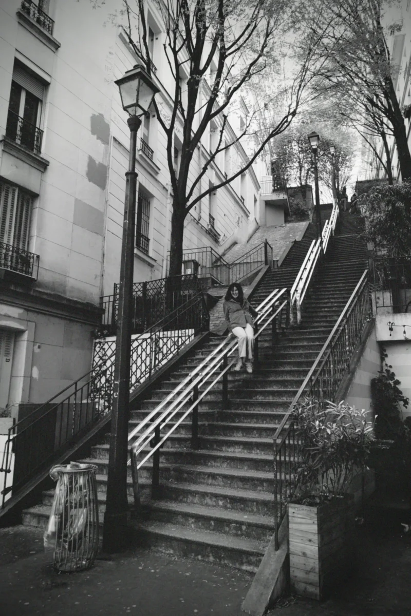 Denis · 22/03/2026 · 3265 x 4898 Cette photographie en noir et blanc met en scène un escalier en pierre bordé de rampes métalliques, sujet principal. La composition est symétrique, avec les marches menant vers un point de fuite.Le cadrage est serré, capturant les détails des bâtiments et des lampadaires. La lumière, douce et naturelle, crée des contrastes subtils, accentuant les textures. Les couleurs dominantes, bien que monochromes, varient en nuances de gris.Le style évoque une esthétique documentaire, typique des clichés urbains classiques. L’ambiance est calme et mélancolique, renforcée par l’absence de mouvement et l’architecture ancienne.