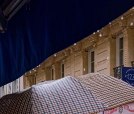 Monique · 17/05/2026 · 4682 x 3360 Cette photographie met en scène un bâtiment classique avec une façade ornée de balcons et de fenêtres symétriques. Le sujet principal semble être un drapeau ou une bâche à carreaux rouges et blancs, partiellement visible en premier plan.La composition joue sur les lignes architecturales et la perspective ascendante. Le cadrage est serré, mettant en valeur les détails de la structure et du drapeau. La lumière, douce et tamisée, crée une ambiance nocturne ou crépusculaire. Les couleurs dominantes sont les tons beiges et blancs du bâtiment, contrastés par les rouges et blancs du drapeau.Le style est sobre et élégant, évoquant une ambiance à la fois historique et légèrement mystérieuse.