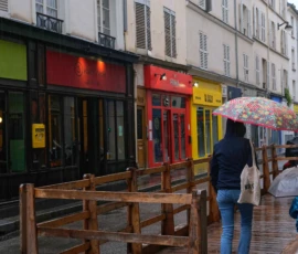 Monique · 20/05/2026 · 5234 x 4160 Cette photographie montre une personne tenant un parapluie coloré sous une légère pluie, marchant sur un trottoir pavé. Le sujet principal est cette personne, dont le parapluie ajoute une touche vibrante.La composition met en valeur les façades colorées des commerces en arrière-plan, avec des tons vifs (vert, rouge, jaune). Le cadrage est serré sur la personne, avec une profondeur de champ qui inclut les enseignes lumineuses. La lumière est tamisée, typique d’un jour gris et pluvieux, créant une ambiance calme et légèrement mélancolique. Les couleurs dominantes sont les teintes vives des enseignes et le parapluie, contrastant avec les tons neutres du pavé et des bâtiments. Le style est réaliste et urbain.