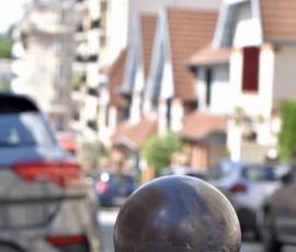 Deniz · 09/05/2026 · 4000 x 6000 Cette photographie met en avant un **boulon de protection routière** en métal, sujet principal flou mais identifiable. La composition joue sur le **cadrage serré** en avant-plan, contrastant avec un arrière-plan flou de rue urbaine.La lumière est naturelle, probablement en journée, avec des ombres douces. Les **couleurs dominantes** sont neutres : gris (boulon, voitures), beige (bâtiments), et touches de vert (végétation). Le style est **documentaire et réaliste**, capturant une scène banale du quotidien.L’ambiance est **calme et urbaine**, avec une impression de mouvement suggéré par les véhicules en arrière-plan.